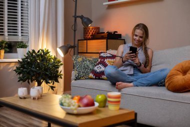 Kanepede dinlenen, abur cuburlardan zevk alan ve akıllı telefonunu kullanan genç bir kadının sakin bir iç mekan sahnesi..