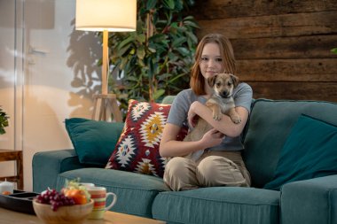 Hoş ve sıcak bir oturma odası, sergilenen vitrinler, zarif kanepesinde küçük şirin bir köpek yavrusu tutan bir kadın.