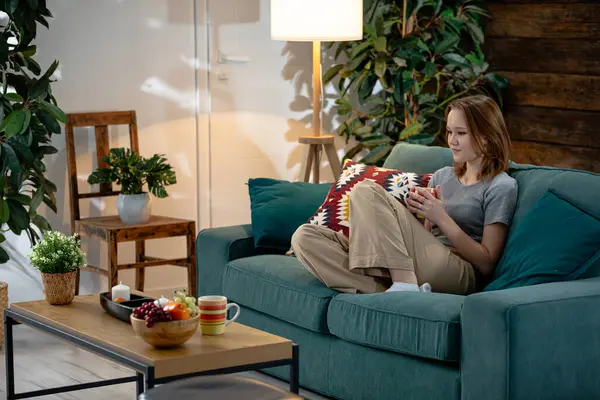 Huzurlu bir oturma odası sahnesi. Dinlenme zamanlarında atıştırmalıkların tadını çıkaran genç bir kadın.