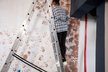 Uzun bir merdivendeki çalışkan bir insan, canlı el izleriyle donatılmış dokulu bir duvarı ustalıkla boyar.