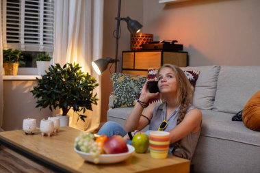 Genç bir kadın evinde rahatça dinleniyor, mutlu bir şekilde abur cubur yiyor ve rahat bir ortamda telefonuyla sohbet ediyor.