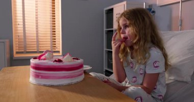 Renkli pijamalı bir kız iyileşme sürecinde hastane odasında pastayla sevinçle kutluyor.