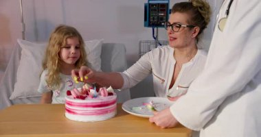 Doğum gününü sevinçle kutlayan bir kız hastane odasında, şefkatli annesi ve dost canlısı bir doktorla birlikte pastayla kutluyor.