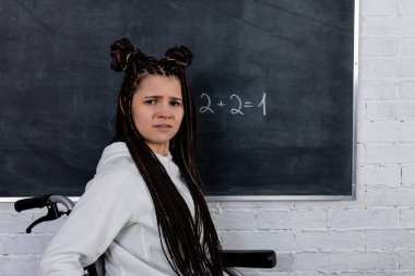 Şık örgüleri olan kendine güvenen genç bir kadın, canlı bir sınıfta tahtayla kameraya bakıyor.