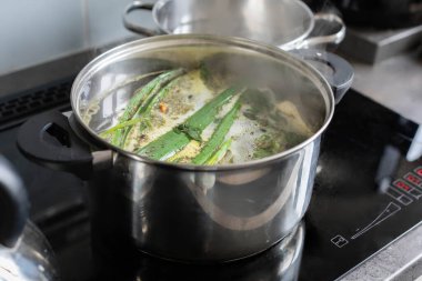 Taze yeşil malzemelerle dolu büyük bir tencere modern ve parlak bir sobanın üstünde buharlaşıyor.