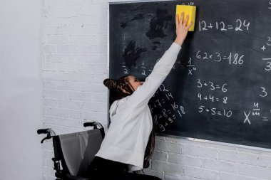 Tekerlekli sandalye kullanan kararlı bir öğrenci matematik denklemleriyle dolu bir tahtayı temizlemek için yaratıcı bir sünger kullanıyor.