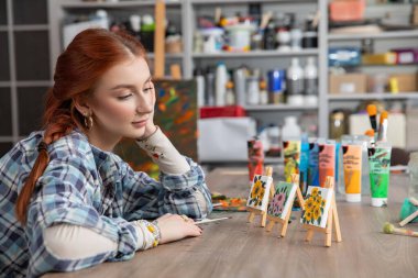 Düşünceli bir sanatçı, rahat stüdyosunda canlı resimler ve küçük sehpalarla yaratıcı bir şekilde çevrilidir.