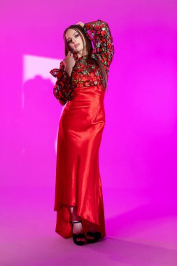 Parlak kırmızı bir elbise giyen göz kamaştırıcı bir manken, saf zarafet ve stil sergileyen pembe bir arkaplanda poz veriyor.