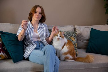 Bir kadın koltukta rahat bir şekilde otururken tüylü, sevimli kedisiyle hoş bir etkileşime girer.