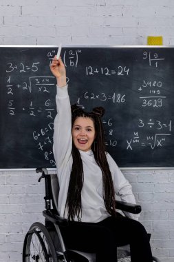 Tekerlekli sandalyedeki neşeli bir öğrenci elini kaldırıyor ve tahtaya karşı matematik başarılarını kutluyor..