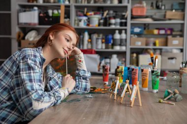 Canlı bir stüdyoda, sanatçının biri düşüncelere dalmış, etrafı renkli resimler ve potensiyelle dolu sehpalarla çevrili.