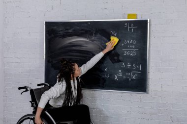 Tekerlekli sandalyedeki dinamik bir öğretmen aktif olarak tahtaya yazı yazıyor, eğitime dahil olmayı teşvik ediyor..