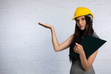 Kasketli genç bir kadın, bir inşaat alanında çalışmalarını sunan bir not panosunu kendine güvenle tutuyor.