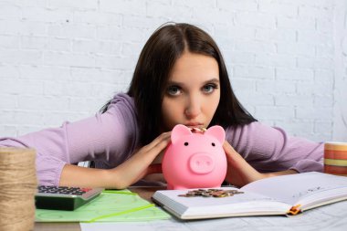 Düşünceli genç bir kadın pembe bir kumbaraya bakıyor, mali durumunu ve gelecek planlarını düşünüyor.