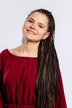 Neşeli genç bir kadın örgülü saçlarıyla poz veriyor, gösterişli bir bordo elbiseyle sade bir arkaplanda.