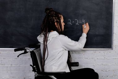 Tekerlekli sandalyedeki kararlı ve kendini işine adamış bir öğretmen öğrencilerini tahtaya yazı yazarak etkilemeye çalışır.