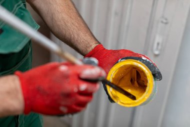 Yetenekli bir işçi konteynırda canlı sarı boyayı karıştırır. Güvenlik ve işçiliği eldivenlerle vurgular.