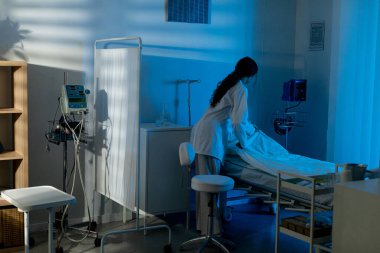 Hemşire bir hastane yatağı ayarlar. Huzurlu, mavi ışıklı bir tıp odasında. Sağlık hizmetleri profesyonelliği gösterilir..