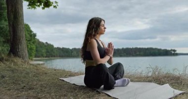 Genç bir kadın, yemyeşil yemyeşil bir gölün kenarında huzur içinde otururken meditasyon yapıyor.