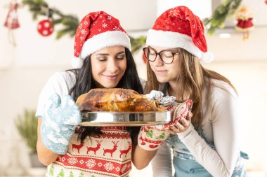 Two cheerful cooks, mom and her teenage daughter, in Christmas aprons with Santa hats, have baked a goose or turkey, they are smelling it. clipart