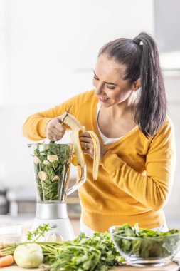 A woman in the kitchen cuts a banana in a blender together with spinach, makes a healthy smoothie.