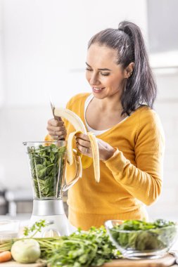 A woman in the kitchen cuts a banana in a blender together with spinach, makes a healthy smoothie.