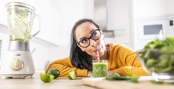 A funny young woman drinks a green smoothie that she made herself.