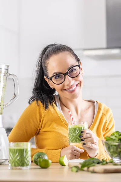 Happy woman with spinach smoothie drink or vegetarian shake sitting in her kitchen.
