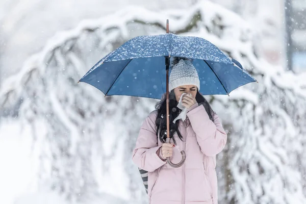 Woman clears her running nose standing on the street under an umbrella on a snowy day.