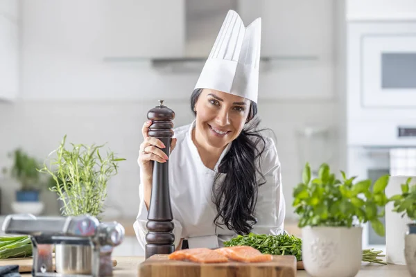 在准备鱼和蔬菜餐的过程中 身穿燕尾服 白帽和制服的女厨师拿着胡椒研磨机 — 图库照片