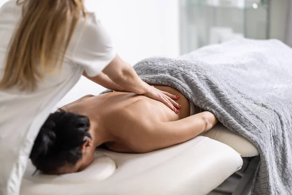 Blonde masseuse gives back massage to a dark-haired client lying face down on massaging table.