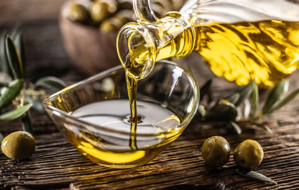 stock image Pouring olive oil from carafe to the glass bowl and in background are olive branches put on table.