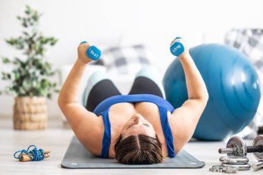 Oturma odasında spor yapan şişman kadın. Paspasa uzanır ve dambılları kaldırır..