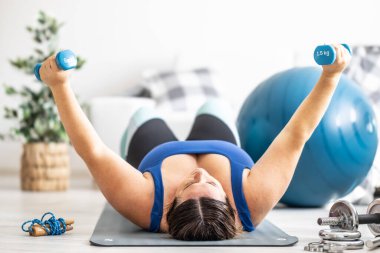 Oturma odasında spor yapan şişman kadın. Paspasa uzanır ve dambılları kaldırır..