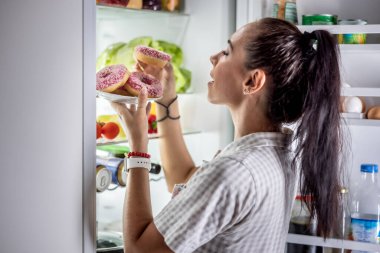 Genç aç bir kadın akşam geç saatlerde açık buzdolabının yanında tatlı çörekler yemek üzere..