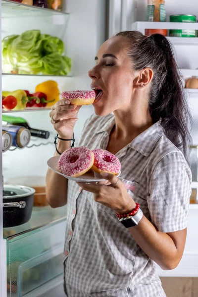穿着睡衣的饥肠辘辘的黑发女郎深夜在开着的冰箱旁享受甜甜的甜甜圈 — 图库照片