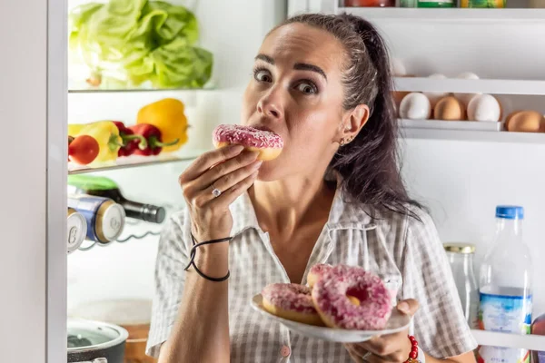 stock image Hungry brunette in pajamas enjoys sweet donuts late at night by the open refrigerator.