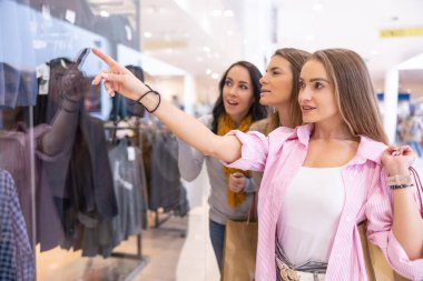 Üç neşeli bayan arkadaş süpermarketin camının arkasındaki kıyafetlere bakıyor..