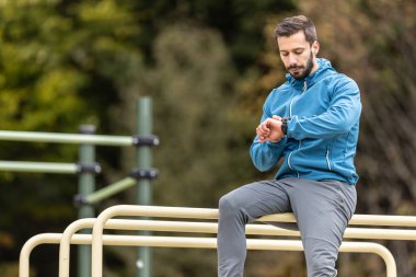Genç bir sporcu doğadaki parmaklıklara oturur ve akıllı bir saat ile hareketlerini izler..