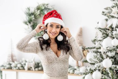 Noel Baba şapkalı kadın Noel ağacı süsleme toplarını tutuyor ve küpeleri kameraya gülümsüyor..