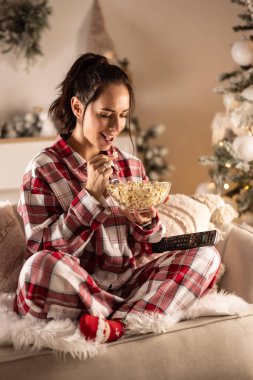 Genç bir esmer, Noel 'de patlamış mısırın tadını çıkarıyor. Pijamalarıyla televizyon karşısında oturuyor..