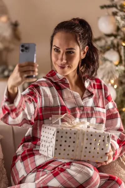 Cheerful woman in pajamas sitting on sofa with gift next to Christmas tree taking selfie.