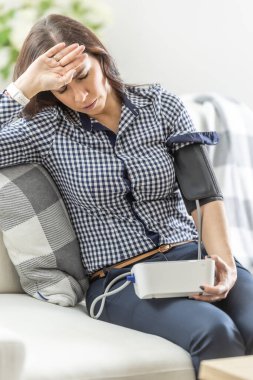 Genç bir kadının baş dönmesi sorunu var, kanepeye oturuyor ve kan basıncını dijital bir ölçüm cihazıyla ölçüyor..