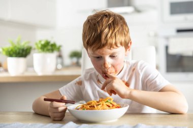 Küçük tatlı bir çocuk öğle yemeği için mutfakta spagetti bolonez yiyor ve ketçapla kaplanmış ve parmaklarını yalıyor..