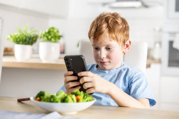 自宅で台所でランチする前にスマートフォンを使用してスマートかわいい男の子の景色 ストック画像