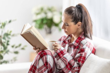 Pijamalı güzel esmer, oturma odasındaki kanepede oturmuş kitap okuyor..