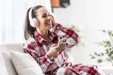 Kanepede oturan gülümseyen genç bir kadın cep telefonuyla kulaklıkla müzik dinliyor..