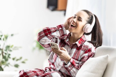 Kanepede oturan gülümseyen genç bir kadın cep telefonuyla kulaklıkla müzik dinliyor..