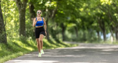 Zinde genç bir kadın parkta koşuyor gülümsüyor mutlu koşuyor ve sağlıklı bir açık hava yaşam tarzının tadını çıkarıyor..