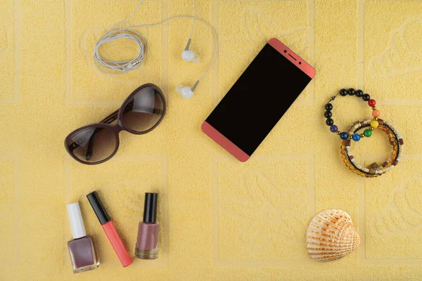 Women\'s accessories for a beach holiday. A smartphone, jewelry and cosmetics, sun glasses, headphones and a shell in the background of a yellow terry towel.